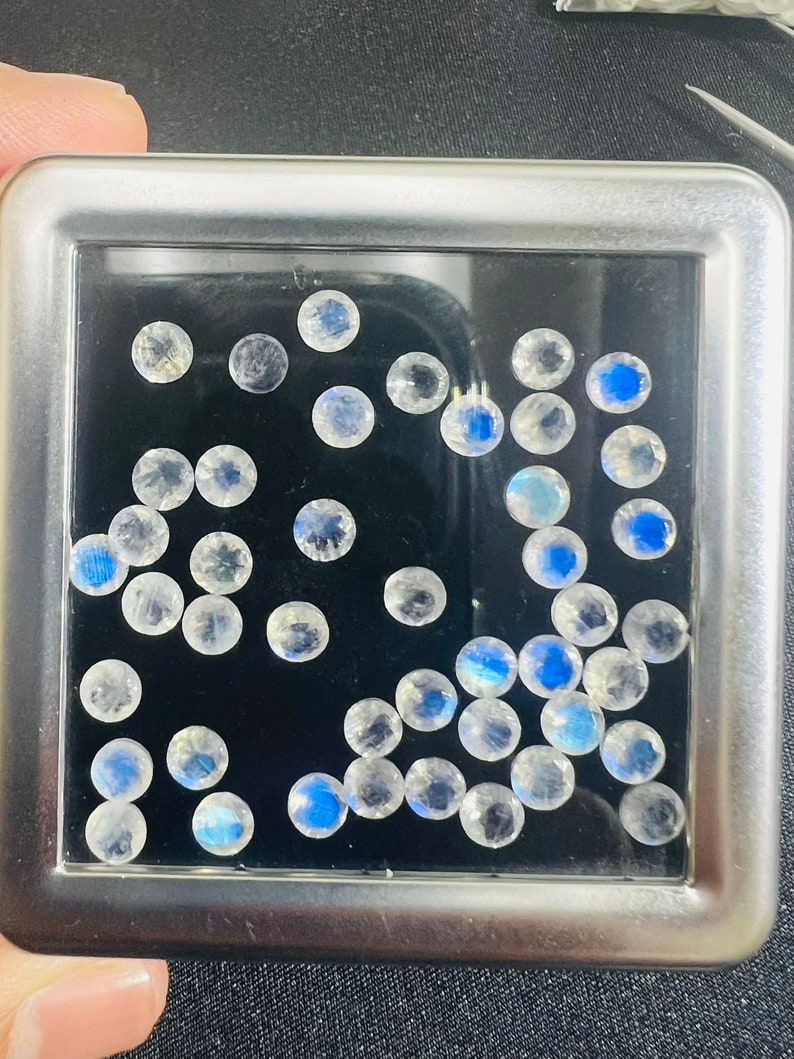 May include: A close-up of a metal box containing a collection of round, iridescent white gemstones. The gemstones are scattered randomly on a black background.
