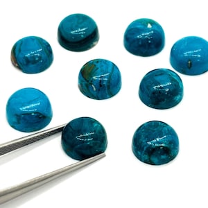 May include: A collection of nine polished, cabochon-cut gemstones in varying shades of teal and turquoise. The stones are round and domed, with unique swirling patterns. A pair of tweezers holds one stone, highlighting its detail.