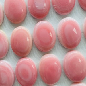 May include: A collection of 15 oval-shaped pink and white shell cabochons. The cabochons are arranged in rows on a white surface.