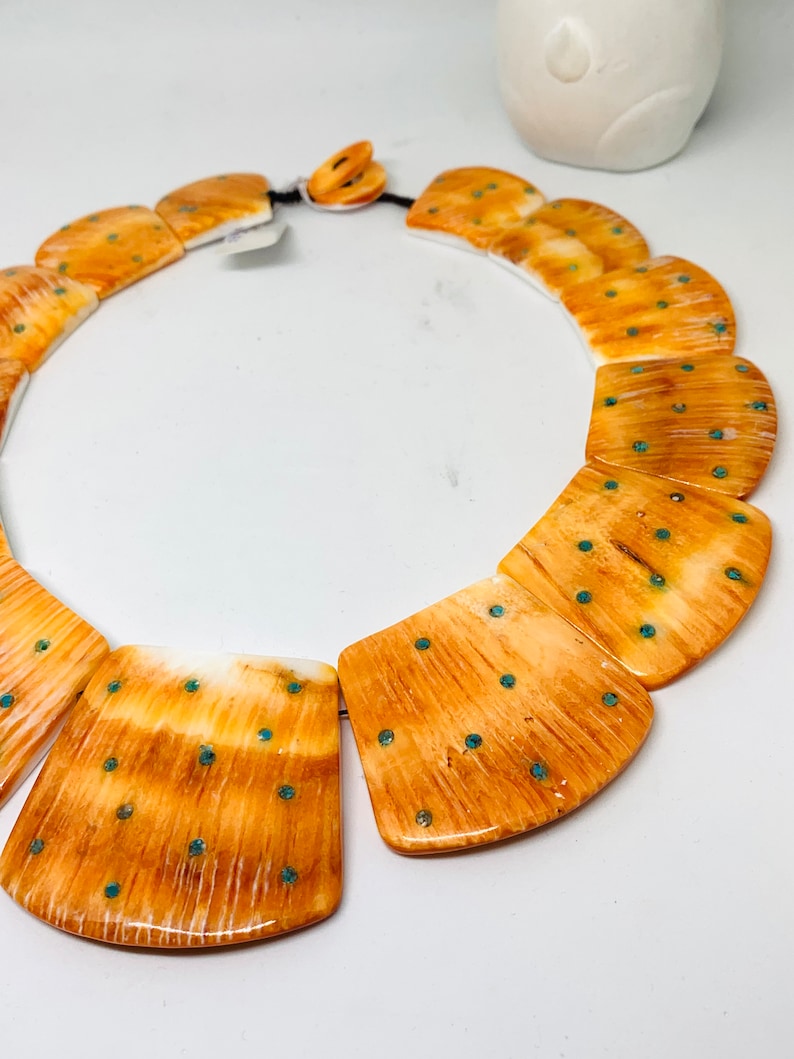 May include: A necklace made of large, rectangular, polished bone beads with a light brown and white marbled pattern. The beads are accented with small, turquoise-colored dots.
