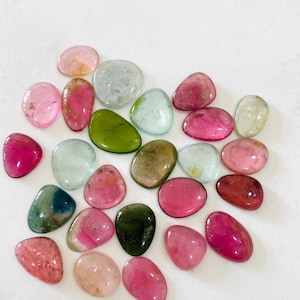 May include: A collection of polished gemstones in various colors, including pink, green, and clear. The stones are oval and rounded, with a smooth, glossy surface. The gemstones are scattered on a white surface.