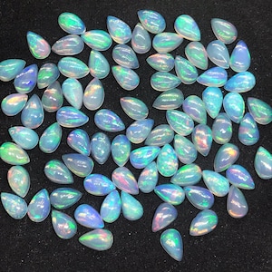 Può includere: Una collezione di pietre preziose di opale a forma di lacrima iridescenti in varie tonalità di blu, verde e rosa. Gli opali sono disposti su una superficie nera.