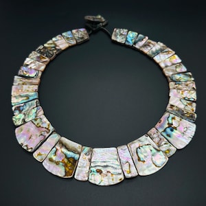 May include: A striking necklace made from iridescent abalone shell segments. The rectangular shell pieces are arranged in a collar, showcasing pink, blue, and gold hues. The necklace is displayed against a black backdrop.