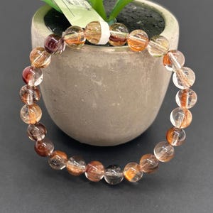 May include: A beaded bracelet featuring clear and amber-colored beads with reddish-brown inclusions. The bracelet is made of round, polished beads strung together. The beads have a translucent quality, allowing light to pass through. The bracelet is displayed near a small potted plant.