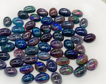 Puede incluir: Una colección de ópalos negros ovalados con colores arcoíris iridiscentes. Los ópalos están dispuestos sobre una superficie blanca.