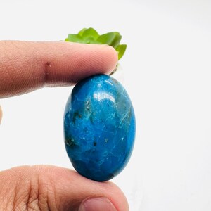 May include: A smooth, oval-shaped blue gemstone with a marbled pattern. The stone is being held by a finger.
