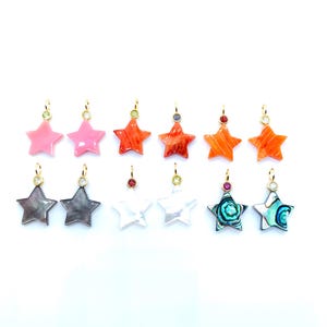 May include: A collection of star-shaped pendants in various colors, including pink, orange, gray, white, and teal. Each pendant has a gold-colored loop and is adorned with a small gemstone. The pendants are displayed against a white background.