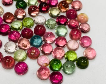 May include: A collection of colorful, round gemstones in shades of pink, green, and clear. The gemstones are scattered on a white surface, with a variety of colors and sizes.