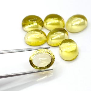 Może przedstawiać: Kolekcja siedmiu złotych, żółtych kamieni szlachetnych w kształcie kaboszonów. Jeden jest trzymany przez pęsetę, odsłaniając blady, półprzezroczysty środek. Pozostałe sześć jest rozproszonych, prezentując gładkie, wypukłe powierzchnie. Kamienie są na jasnym, białym tle.