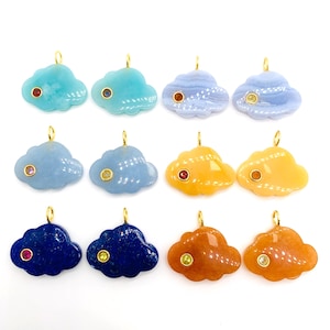 Pode incluir: Uma coleção de pingentes em forma de nuvem em várias cores, incluindo azul, laranja e azul escuro. Cada pingente apresenta uma pequena pedra preciosa colorida e uma argola dourada para pendurar. Os pingentes são exibidos em um fundo branco.