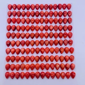 May include: Rows of teardrop-shaped, polished red gemstones arranged on a white surface. The gemstones have a vibrant, reddish-orange hue with natural patterns. The image shows a collection of these gemstones, likely for jewelry making or crafting.