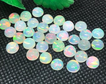 May include: A collection of round, cabochon-cut opals. The opals are mostly white with iridescent flashes of color, including pink, green, and yellow. They are arranged on a black textured surface, with a green leaf in the background.