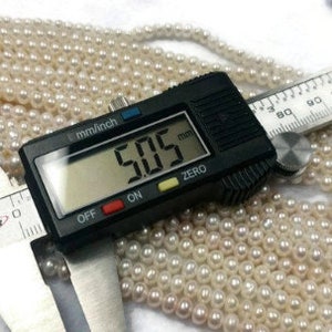 May include: A digital caliper displays a measurement of 5.05 mm, positioned over a collection of small, off-white pearls. The pearls are arranged in multiple strands. A ruler is also visible, with the word "SUPER" on the caliper.
