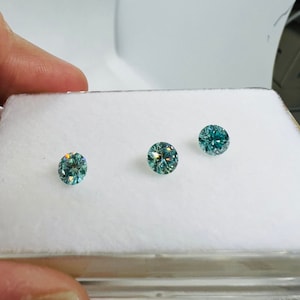 May include: Three round, faceted, teal green gemstones are displayed in a white box. The gemstones are arranged in a row.