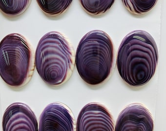 May include: Twelve oval-shaped cabochons with a polished, glossy finish. The cabochons feature concentric, swirling patterns in shades of purple, white, and burgundy. The dimensions are 18 x 25mm. The cabochons are arranged on a white surface.