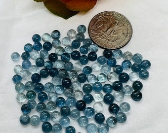 May include: A collection of approximately 60 round, light blue aquamarine gemstones. The stones are smooth and polished, and they are arranged in a loose pile on a white surface. A US quarter is included for scale.