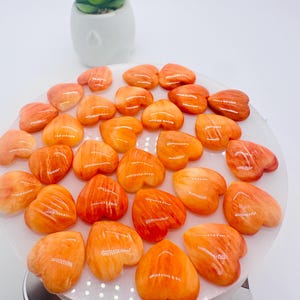 May include: A collection of 20 heart-shaped orange and white striped stones. The stones are arranged in a circle on a white surface.