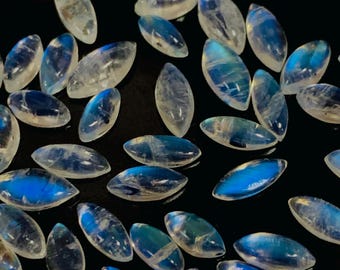Puede incluir: Colección de piedras preciosas de forma ovalada, piedras de luna con un brillo iridiscente azul y blanco. Las piedras están dispersas sobre una superficie negra, mostrando su aspecto pulido y translúcido. Ideal para joyería.