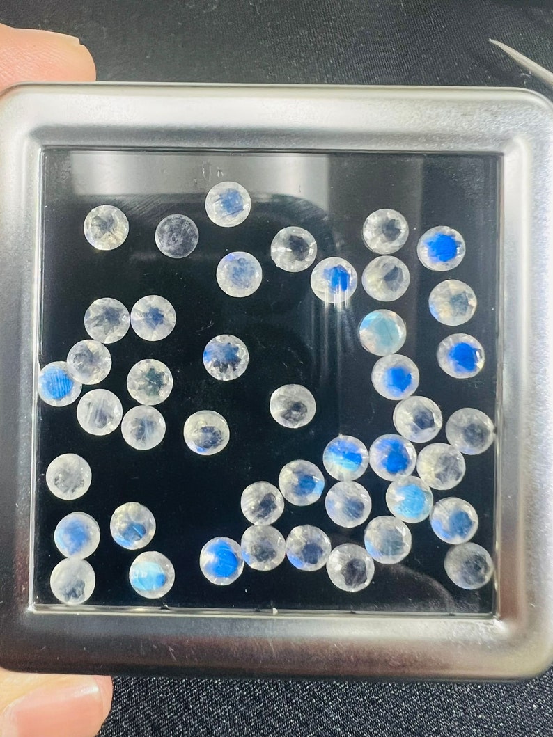 May include: A collection of round, iridescent moonstone gemstones in a clear plastic container. The stones are various sizes and have a milky white color with a blue sheen.