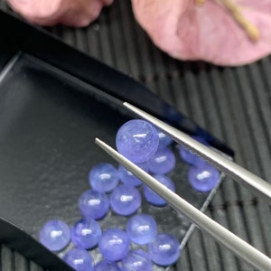 May include: A close-up of a collection of round, light blue gemstones in a black tray. The gemstones are arranged in a semi-circle pattern. A pair of tweezers holds one of the gemstones.