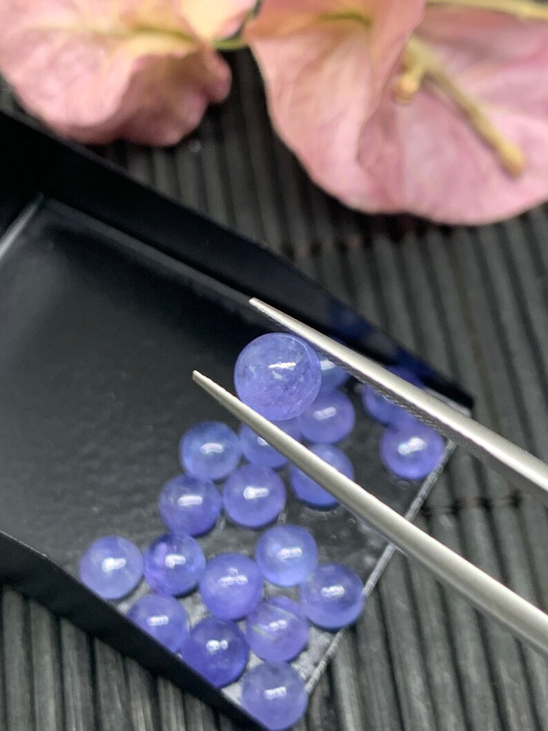 May include: A close-up of a collection of round, light blue gemstones in a black tray. The gemstones are arranged in a semi-circle pattern. A pair of tweezers holds one of the gemstones.
