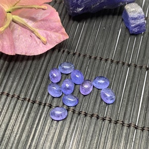 May include: A group of oval-shaped, light blue gemstones on a dark brown woven mat. The gemstones are smooth and translucent.