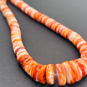 May include: A necklace made of orange and white shell beads. The beads are flat, round, and strung together. The necklace is displayed on a dark gray surface.