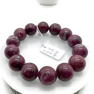 May include: A purple gemstone bracelet with 12 round beads. The bracelet is on a white display stand with a black and white pattern. A small tag is attached to the bracelet with the text "8.9g 101.0 EA".