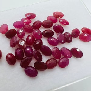 May include: A collection of oval-shaped, faceted ruby gemstones in various shades of pink and red. The gemstones are arranged in a loose pile on a white surface.
