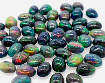 Puede incluir: Una colección de ópalos negros de forma ovalada con destellos iridiscentes verdes, azules y rojos. Los ópalos están esparcidos sobre una superficie blanca.