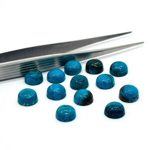 May include: A collection of turquoise cabochon gemstones, each with a domed shape and varying shades of blue and green. A pair of silver tweezers is positioned near the gemstones, suggesting their use in jewelry making or lapidary work.