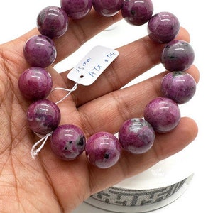 May include: A pink and purple gemstone bracelet with 15mm beads. The bracelet is on a person's wrist and has a tag that says "15mm ATX # D4".