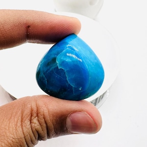 May include: A teardrop-shaped blue gemstone with white and green veins. The gemstone is being held in a hand.