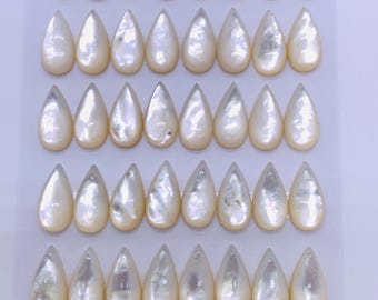 Könnte beinhalten: Eine Anordnung von 40 tropfenförmigen, schillernden, cremefarbenen Perlmutt-Cabochons, jeweils 8x16mm groß, auf einer weißen Karte. Die Cabochons haben einen glänzenden, perlmuttartigen Schimmer.