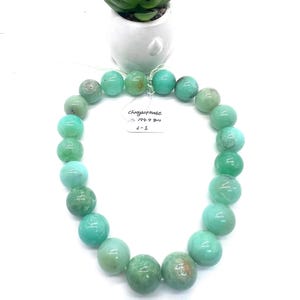 May include: A necklace made of large, round, light green chrysoprase beads. The beads are strung together, forming a circular shape. A small white tag with handwritten text is attached. A small potted succulent plant sits above the necklace.