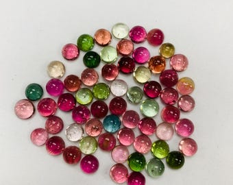 May include: A collection of polished, round gemstones in various colors, including pink, green, red, and clear. The gemstones are scattered on a white surface, showcasing their translucent quality and diverse hues. These stones are likely used for jewelry making.