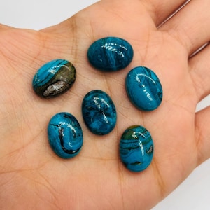 May include: Six oval-shaped gemstones in shades of teal and turquoise are displayed. The stones have unique patterns of dark blue, brown, and black veining. The gemstones are smooth and polished, with a glossy finish.