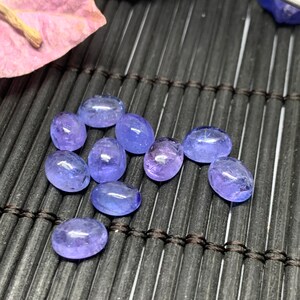 May include: A group of ten oval-shaped, translucent blue gemstones on a black woven mat. The gemstones have a light blue color and are smooth and polished.