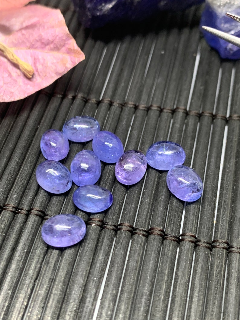 May include: A group of ten oval-shaped, translucent blue gemstones on a black woven mat. The gemstones have a light blue color and are smooth and polished.