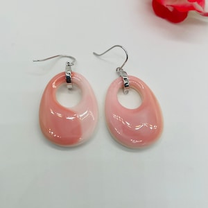 May include: A pair of dangle earrings with a light pink and white oval design. The earrings have a silver-colored hook and a small silver-colored ring connecting the hook to the oval. The earrings are on a white background.