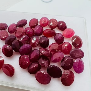 May include: A collection of oval-shaped, faceted ruby gemstones in various shades of red and pink. The gemstones are displayed in a clear plastic tray on a white surface.