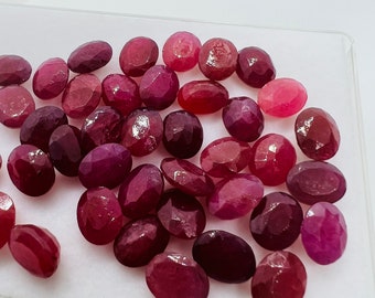 Könnte beinhalten: Eine Sammlung ovaler, facettierter Rubin-Edelsteine in verschiedenen Rot- und Rosatönen. Die Edelsteine werden in einer durchsichtigen Plastikschale auf einer weißen Oberfläche präsentiert.