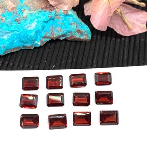 Puede incluir: Doce piedras preciosas rectangulares rojas, probablemente granates, dispuestas en tres filas de cuatro sobre una superficie blanca.