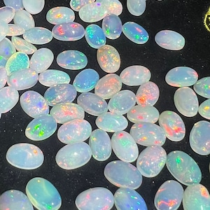 May include: A collection of oval-shaped white opals with iridescent rainbow colors. The opals are scattered on a black surface.