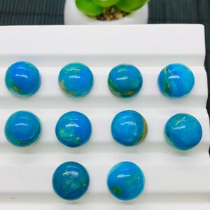 May include: Ten round, blue and green cabochon gemstones arranged in three rows on a white surface.