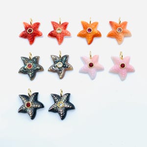 May include: Ten starfish-shaped pendants arranged in three rows. Each pendant features a textured surface, a gold-tone metal bail at the top, and a small circular gemstone set in the center. The colors include shades of red, orange, pink, and dark gray.
