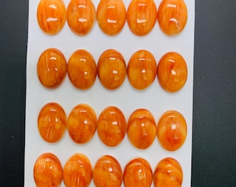 Könnte beinhalten: Zwanzig ovale, polierte Edelsteine in Orange- und Bernsteintönen, angeordnet auf einer weißen Karte. Die Maße betragen 13 x 18 mm. Die Edelsteine haben eine glänzende Oberfläche und ein natürliches, marmoriertes Aussehen.