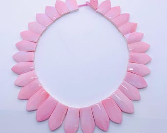 May include: A pink statement necklace featuring petal-shaped beads. The beads are a gradient of light to medium pink, strung together with a pink cord. The necklace is displayed on a white background.