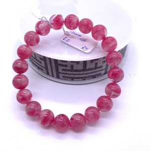 May include: A pink and white gemstone bracelet with 10mm beads. The bracelet is on a white surface with a roll of black and white patterned tape.