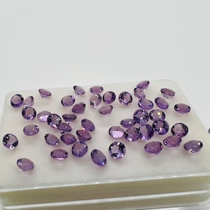 May include: A collection of loose amethyst gemstones in a clear plastic container. The gemstones are oval shaped and have a light purple hue.